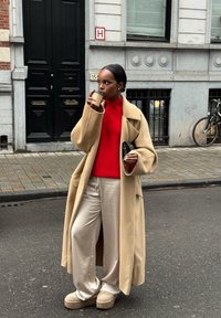 Beżowy oversize płaszcz, czerwony prążkowany sweter, beżowe satynowe spodnie z szerokimi nogawkami oraz buty na platformie. Trzyma kubek z kawą, stoi na ulicy.