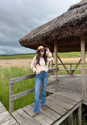 Blusa beige de manga larga y jeans acampanados azules, combinados con un sombrero marrón claro y gafas de sol. De pie sobre una plataforma de madera con techo de paja.
