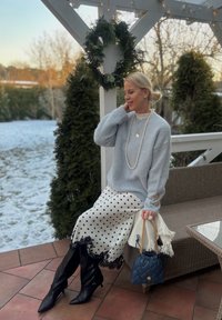 Femme en pull bleu clair et jupe à pois assise sur un banc en plein air, tenant un sac bleu marine, avec un sol enneigé et de la verdure en arrière-plan.