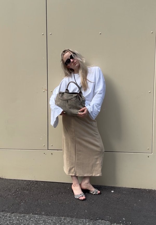 Kobieta ubrana w oversize'ową białą bluzkę, beżową midi spódnicę, czarne okulary przeciwsłoneczne, sandały w zebra print, trzymająca szarą torebkę, stoi na tle beżowej ściany.