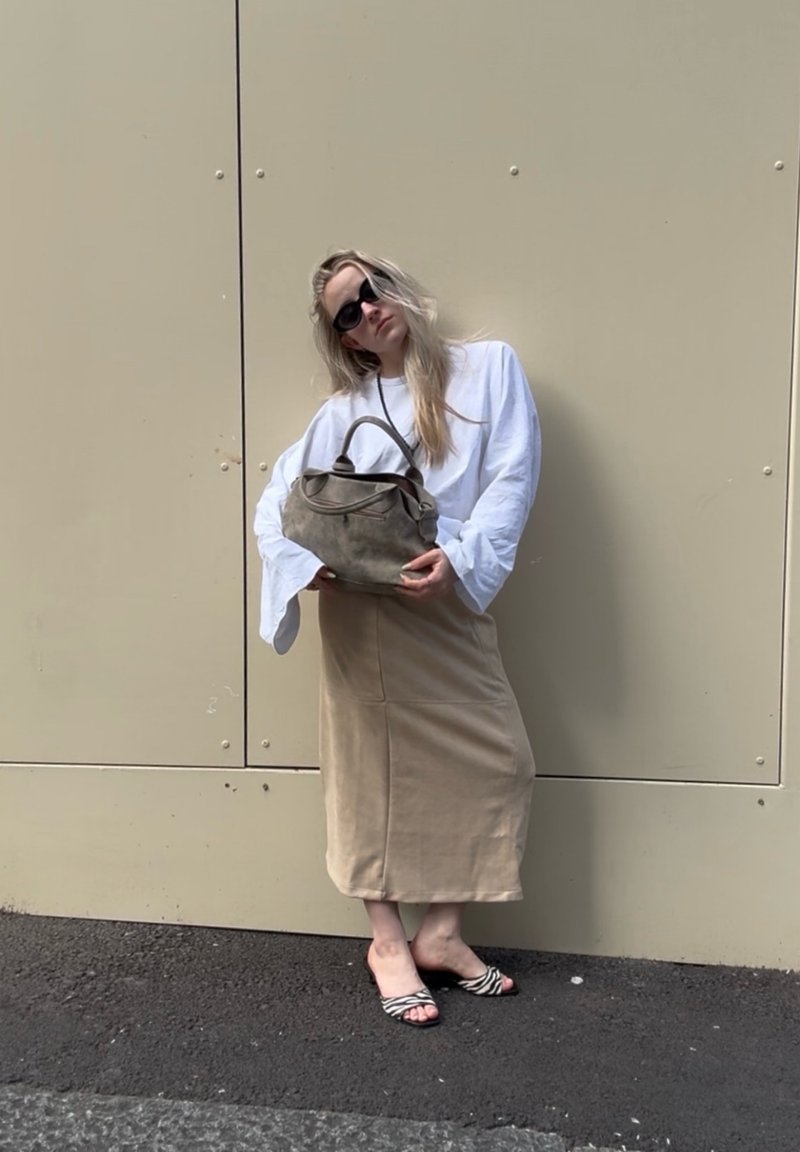 Γυναίκα με λευκό φαρδύ πουκάμισο, μπεζ μίντι φούστα, μαύρα γυαλιά ηλίου, σανδάλια με σχέδιο ζέβρας, κρατώντας γκρι τσάντα, στέκεται απέναντι σε μπεζ τοίχο.