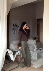 Pulover maro cu blană, pantaloni largi verzi, ținând o geantă maro. Interior modern vizibil cu o canapea albă și o masă de marmură.