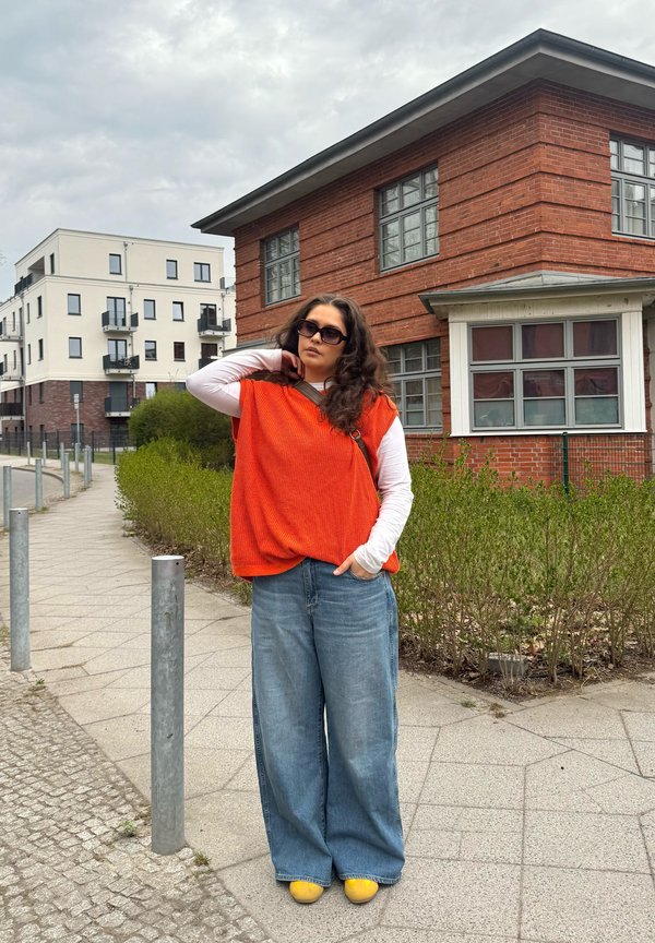 Femme portant un pull sans manches orange et un jean large se tient sur le trottoir près de bâtiments résidentiels modernes sous un ciel nuageux.