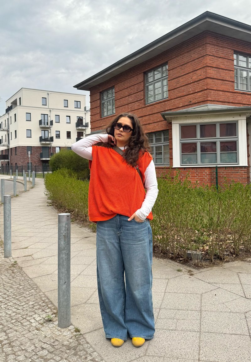 Femme portant un pull sans manches orange et un jean large se tient sur le trottoir près de bâtiments résidentiels modernes sous un ciel nuageux.