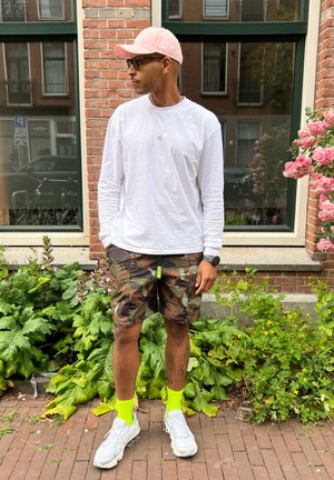 Camisa blanca de manga larga, pantalones cortos de camuflaje, gorra rosa, calcetas verde neón y zapatillas grises. De pie contra una pared de ladrillo con vegetación y flores.