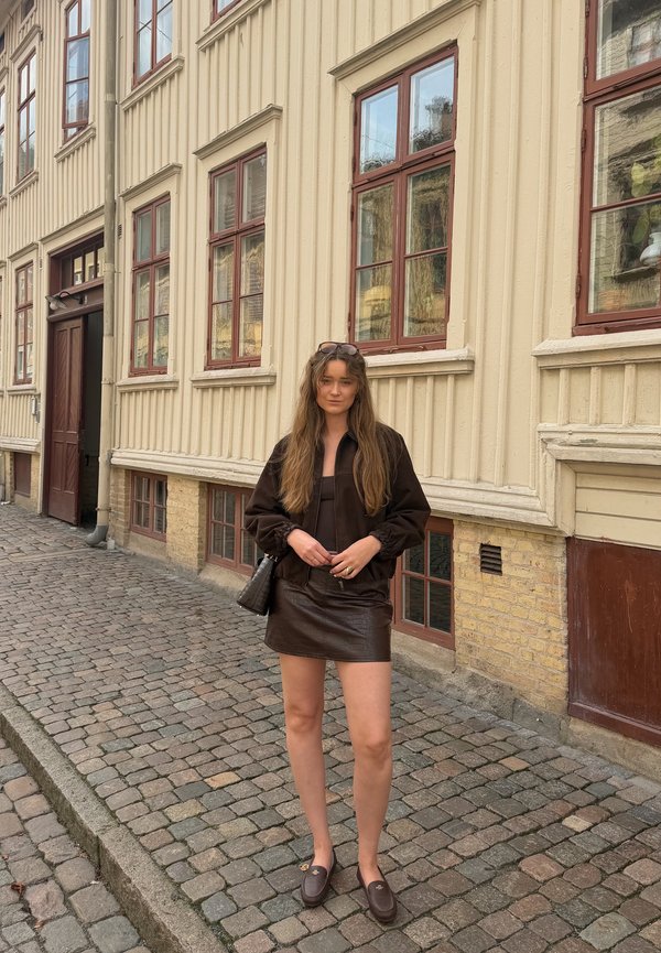 Joven mujer con cabello largo, con chaqueta oscura y falda, está de pie en una calle empedrada cerca de un edificio de color crema con grandes ventanas.