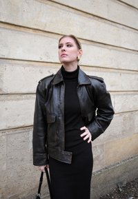 Ruskea nahkatakki, jossa on hartiapaikat, etutaskut ja vetoketjukiinnitys, käytetty mustan ribbikuitupaidan mekon päällä. Yksivärinen, sileä pinta.