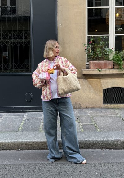 Chaqueta acolchada con estampado floral y ribete rosa, camiseta blanca, jeans de mezclilla azul de corte oversize y bolso de mano beige texturizado. Sosteniendo un pastelito.