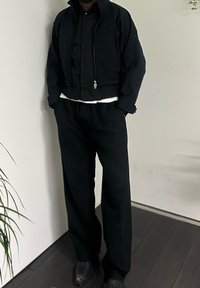 Veste noire courte avec un col montant et une fermeture éclair, associée à un pantalon noir ample. La tenue est superposée sur une chemise blanche.