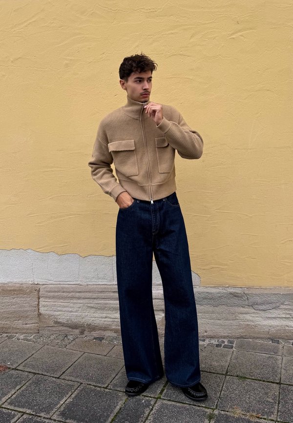 Un homme pose devant un mur jaune, vêtu d’un pull beige zippé, d’un jean foncé à jambes larges et de chaussures noires.