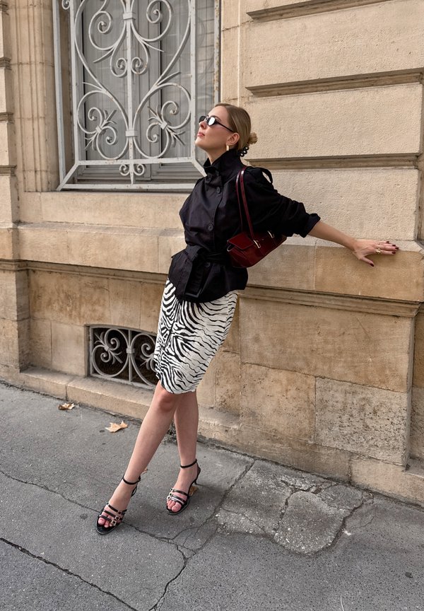 Una mujer con gafas de sol posa junto a un edificio de piedra. Lleva una chaqueta negra, una falda de estampado de cebra, sandalias y un bolso rojo al hombro.