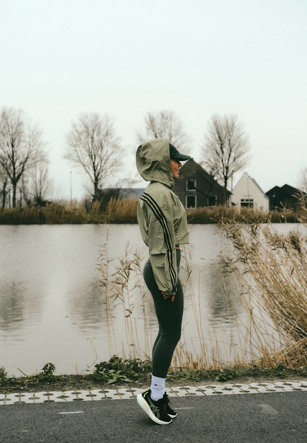 Olijfgroen cropped jack met een capuchon, zwarte strepen en aansluitende donkere leggings. Zwarte sportschoenen met witte accenten. Kalme waterachtergrond.