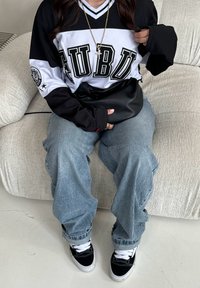 Μαύρη και λευκή μπλούζα με μακρυσά μανίκια και λογότυπο "FUBU", συνδυασμένη με ανοιχτό μπλε φαρδύ τζιν και μαύρα αθλητικά παπούτσια. Τα αξεσουάρ περιλαμβάνουν μια χρυσή αλυσίδα.