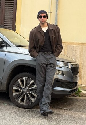 Joven con gafas de sol, chaqueta marrón, gorro negro y pantalones grises, apoyado de manera casual contra un coche gris en una calle de la ciudad.