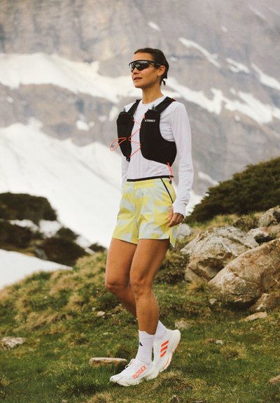 Conjunto deportivo que incluye un chaleco de hidratación negro, una camiseta de manga larga blanca y pantalones cortos con un patrón geométrico en amarillo. Zapatillas deportivas con detalles en naranja.