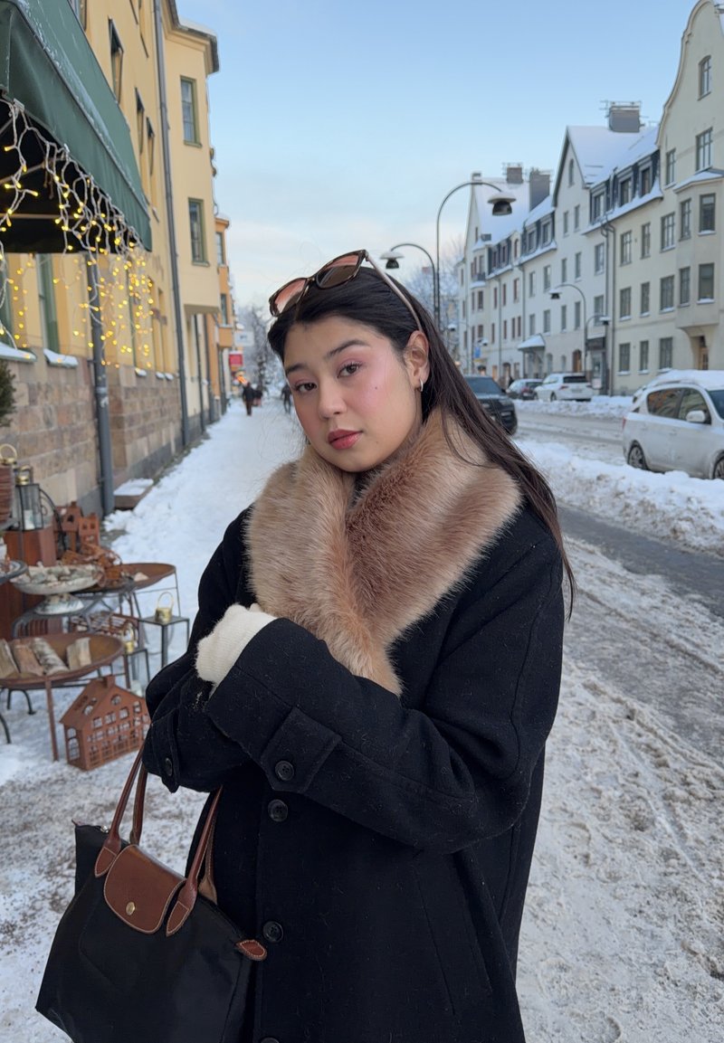 Jonge vrouw in een zwarte jas met bontkraag en handschoenen staat op een besneeuwde straat met gebouwen en geparkeerde auto's, terwijl ze een zwarte handtas vasthoudt.