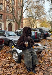 Personne en veste à capuche en fourrure marron et pantalon camouflage tenant un café glacé, assise sur une petite remorque entourée de feuilles d'automne dans un parking urbain.