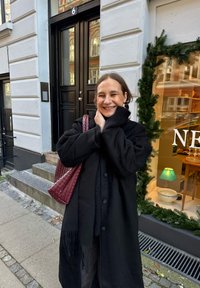 Black coat with textured sleeves, purple woven handbag, black scarf, standing outside a building with a glass door and decorative pine.
