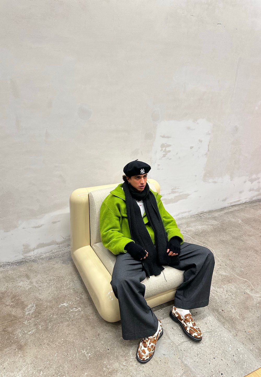 Πράσινη φλις ζακέτα, μαύρο κασκόλ, γκρι παντελόνι σε φαρδιά γραμμή, καφέ και λευκά παπούτσια με σχέδιο, καθισμένος σε έναν κρεμ, γλυπτικό καναπέ απέναντι από έναν απλό τοίχο.