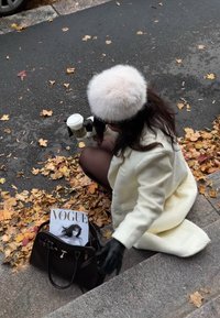 Cappotto di lana bianco, guanti neri e cappello di pelliccia. Borsa nera con una rivista a terra. Foglie autunnali circondano la scena. Tazza di caffè in mano.