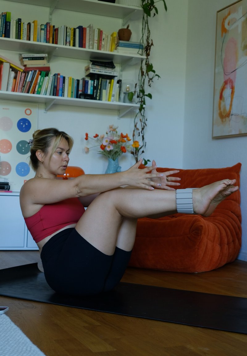 Femme en haut rouge et short noir faisant de l'exercice sur un tapis, en équilibre sur le dos avec les jambes et les bras tendus dans un salon à la maison.