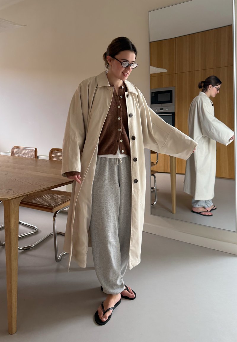 Femme portant un manteau beige, un cardigan marron boutonné, un pantalon de survêtement gris et des tongs noires, debout près d'une table en bois et d'un grand miroir.