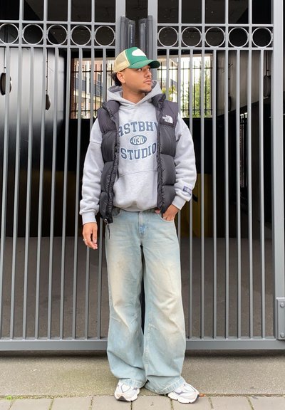 Chaleco acolchado negro sobre sudadera gris con el texto "ASTBH STUDIO", jeans holgados azul claro y zapatillas blancas. Gorro verde y blanco. Fondo de una puerta de metal.