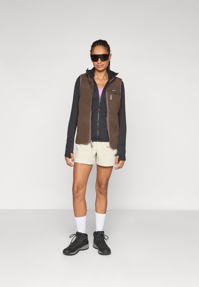 Chaleco de forro polar marrón sobre una sudadera negra con cremallera, combinado con pantalones cortos de color beige claro, calcetines blancos y botas de senderismo negras. Las gafas de sol completan el look.
