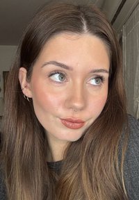 Nuori nainen, jolla on pitkät ruskeat hiukset, vihreät silmät, kiiltävät huulet, nenäkoru ja useita korvien lävistyksiä, katsoo hieman sivulle sisätiloissa.