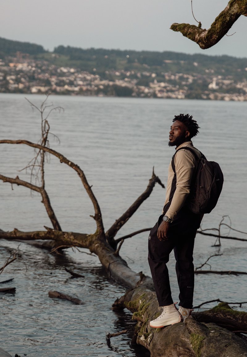 Tenue décontractée avec un haut beige à manches longues et un pantalon noir. Bottes blanches avec des accents rouges et un sac à dos noir. Cadre naturel près de l'eau.