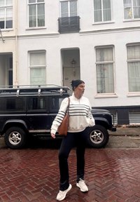 White zip-up hoodie with black stripes, black leggings, and white sneakers. Brown handbag. Black SUV parked on a brick street with white buildings.