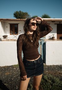 Γυναίκα με γυαλιά ηλίου που σκιάζει τα μάτια της με το χέρι, φοράει καφέ μπλούζα με μακριά μανίκια και τζιν φούστα, στέκεται έξω δίπλα σε λευκό τοίχο και σπίτι.