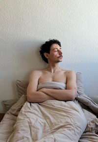 Ανοιχτό μπεζ σεντόνια σκεπάζουν ένα άτομο με σγουρά μαλλιά, που έχει τα χέρια του σταυρωμένα. Ένας μινιμαλιστικός τοίχος χρησιμεύει ως φόντο.