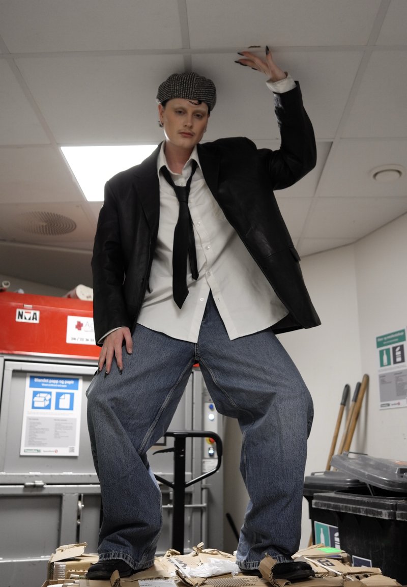 Personne portant un blazer noir, une chemise blanche, un jean ample et une casquette à motifs, posant debout sur des cartons empilés dans un cadre industriel.