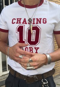 White t-shirt with red trim and "CHASING 10" text, layered silver chains with a pendant, multiple silver rings, decorative bracelet, and a belt.