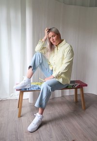 Helles gelbes Hemd mit langen Ärmeln, kombiniert mit hellblauen Jeans und weißen High-Top-Sneakern, sitzt auf einer bunten Bank.