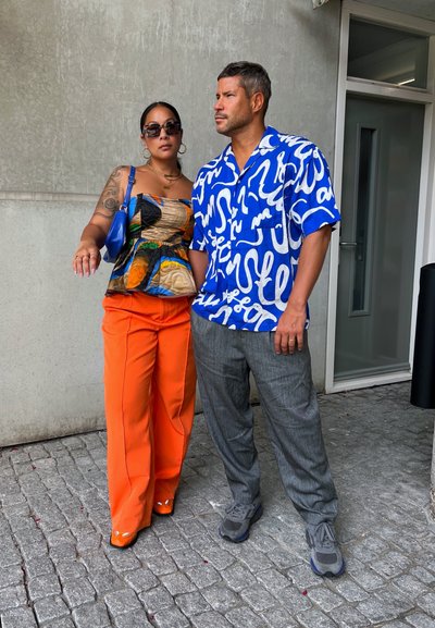 Dos personas están juntas. La mujer lleva un top sin mangas con un patrón y pantalones anchos naranjas. El hombre viste una camisa azul brillante y pantalones grises.