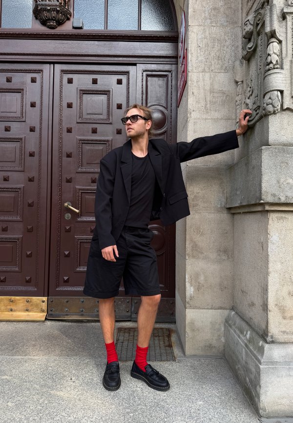 Uomo con blazer nero, pantaloncini, calzini rossi e occhiali da sole che si appoggia a una colonna di pietra vicino a grandi porte di legno marrone.