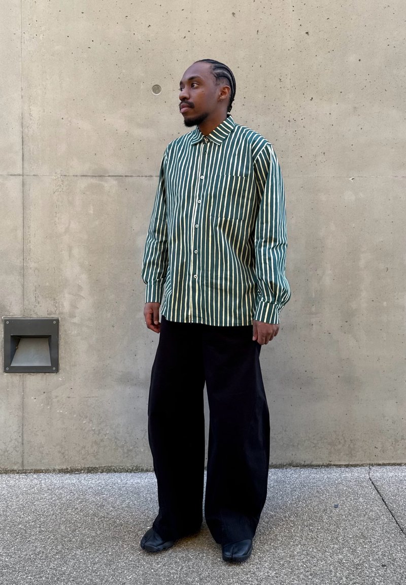 Homme aux cheveux tressés portant une chemise rayée verticalement verte et blanche, un pantalon large noir et des chaussures noires, debout contre un mur en béton.