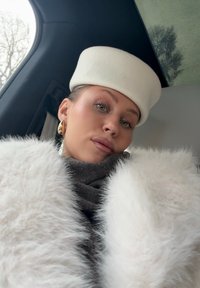 Femme portant un manteau en fausse fourrure blanche, une écharpe grise, de grandes boucles d'oreilles en or et un chapeau blanc rond, assise à l'intérieur d'un véhicule près d'une fenêtre.