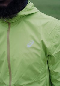 Lys grønn jakke med glidelås foran, med en reflekterende logo på venstre bryst. Materialet virker glatt og vindtett.
