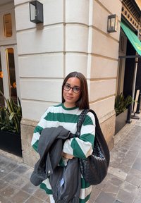 Jeune femme portant des lunettes, vêtue d'une chemise rayée verte et blanche, tenant un manteau et un sac à main en tissu noir tissé, debout au coin d'un bâtiment.