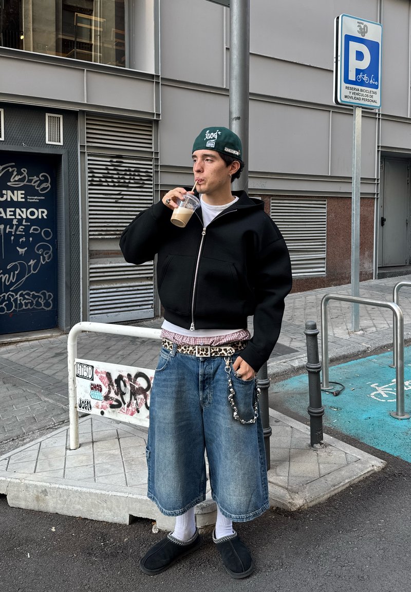 Black zip-up hoodie, white shirt, plaid belt, oversized denim shorts, silver chain, white socks, black slip-on shoes, holding iced coffee.