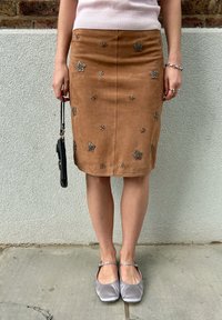 Bruine suède pencilrok met bloemen- en sterversieringen, kniehoogte, gecombineerd met grijze flats, vastgehouden met een kleine zwarte tas.