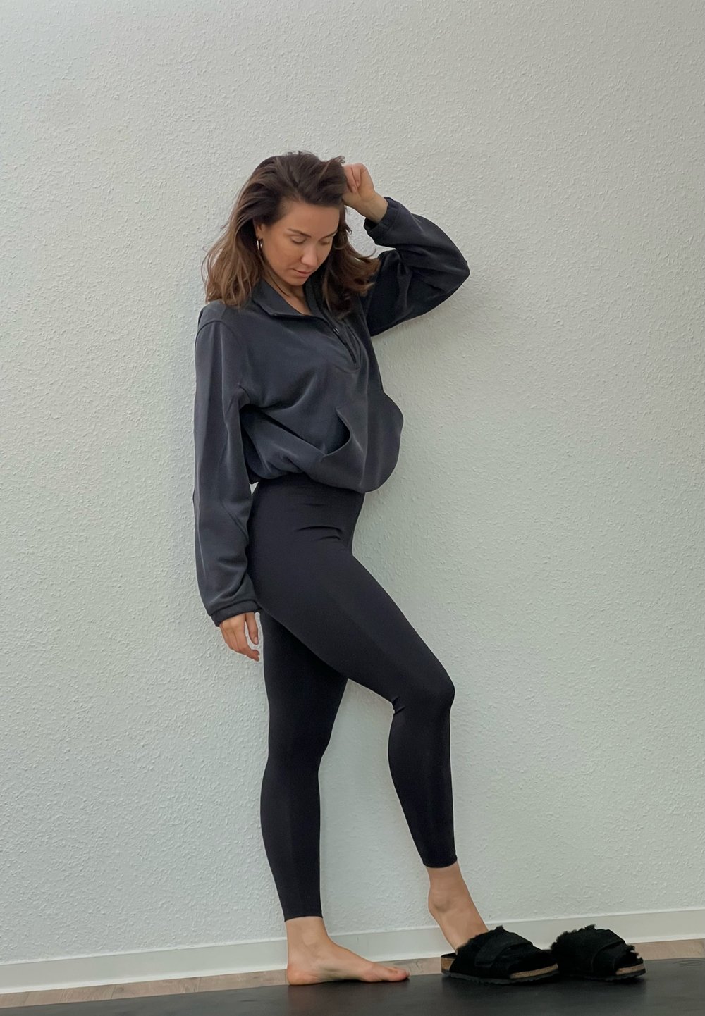 Black zip-up hoodie and leggings, both made from a smooth, stretchy fabric; open-toed black fluffy slippers; light wall background.