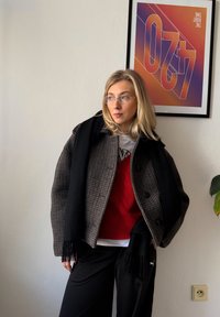 Manteau à carreaux gris avec de gros boutons, écharpe noire, pull rouge en maille torsadée, et pantalon noir. En arrière-plan, une œuvre d'art géométrique.