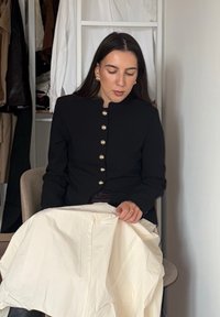 Veste noire structurée avec des boutons dorés, portée sur une jupe plissée couleur crème. Texture lisse, assise sur une chaise simple.