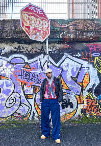 Un hombre se encuentra junto a una pared cubierta de graffiti, viste un suéter de patrón colorido, una chaqueta de cuero negra, pantalones azules sueltos y zapatos beige.