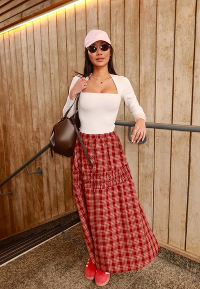 Mujer con top blanco de escote cuadrado, falda larga de cuadros rojos, gorra rosa, gafas de sol, zapatillas rojas y bolso marrón, en unas escaleras junto a una pared de madera.