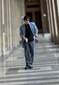 Heled sinise ülikonna jakk musta T-särgi peal, koos laialdaste hallide teksade ja mustade tunkedega. Hoidab valget kruusi.